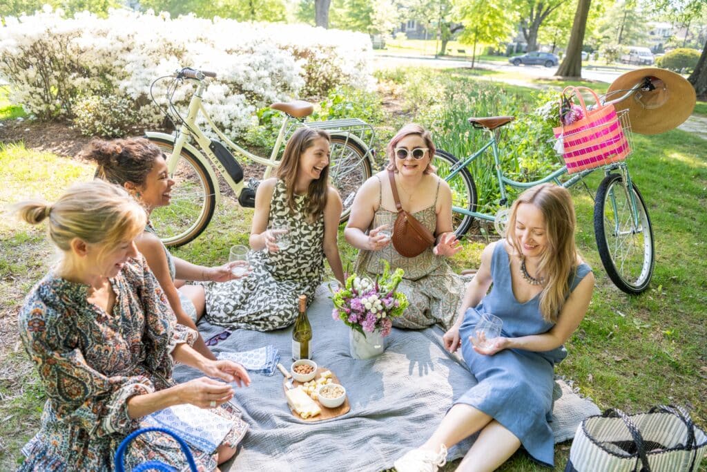 A group of women sit on a patch of grass with bikes around them. They are having a picnic in beautiful spring dresses and are smiling.